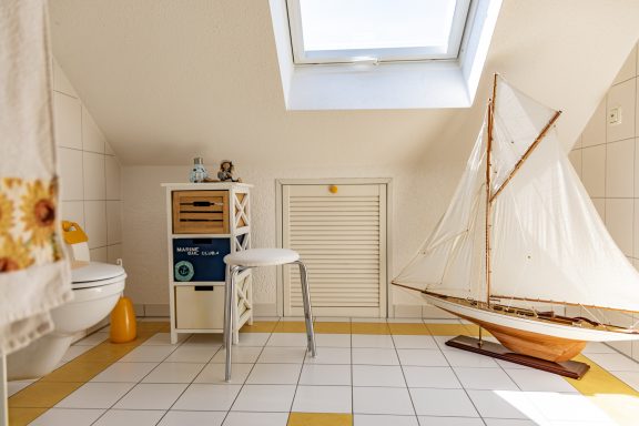 Heller Raum mit Dachfenster, einer weißen Toilette, einem Stuhl und einem Segelboot.
